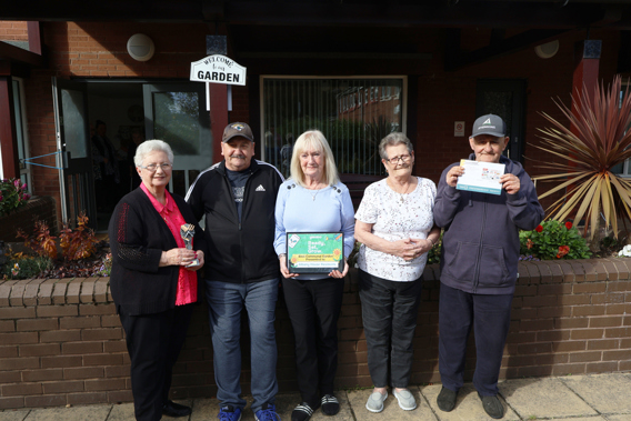 Residents of Albany House Gardening club standing beside the garden holding a trophy, certificate and vouchers